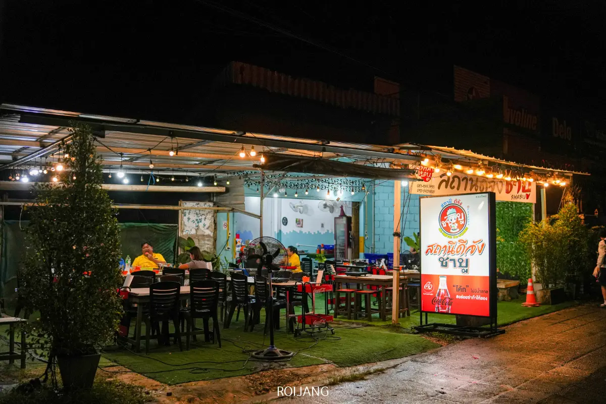 ร้านอาหารไทยกลางแจ้งในเวลากลางคืน มีลูกค้าหลายท่านนั่งอยู่ที่โต๊ะใต้แสงไฟประดับ ไฟนีออนสว่างไสวพร้อมข้อความภาษาไทย รวมถึงโลโก้ สถานีดีจัง แสดงอยู่ที่ทางเข้า.