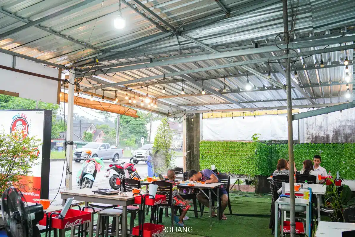 ร้านอาหารกลางแจ้ง สถานีดีจัง มีหลังคาเหล็ก เก้าอี้สีแดง และมีผู้คนหลายคนที่กำลังรับประทานอาหารที่โต๊ะ; มีรถจักรยานยนต์และถนนมองเห็นได้จากภายนอก.