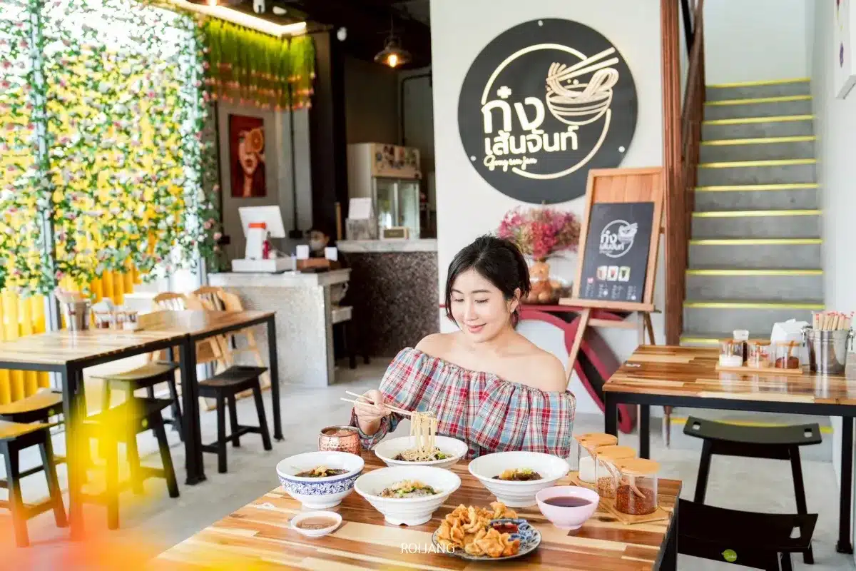 ผู้หญิงคนหนึ่งนั่งอยู่ที่โต๊ะในร้านอาหารสมัยใหม่ ยิ้มและใช้ตะเกียบกินก๋วยเตี๋ยวผัดแห้ง อาหารหลากหลายจานวางกระจายอยู่ตรงหน้าเธอ โดยมีป้ายร้านขนาดใหญ่ติดอยู่บนผนังด้านหลัง.