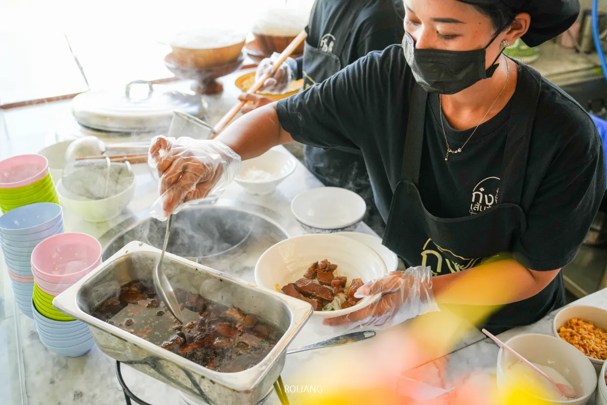 บุคคลที่สวมถุงมือและหน้ากากกำลังตักเนื้อตุ๋นใส่ชามที่เคาน์เตอร์อาหาร ก๋งเส้นจันท์ โดยมีชามสีสันสดใสวางซ้อนกันอยู่ใกล้ๆ.