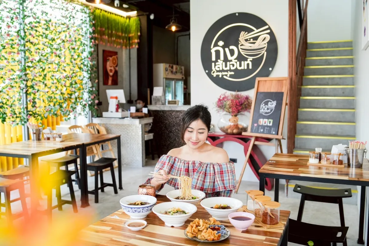ผู้หญิงคนหนึ่งนั่งอยู่คนเดียวที่โต๊ะในร้านอาหารสมัยใหม่ กำลังเพลิดเพลินกับก๋วยเตี๋ยวเส้นจันท์ท่ามกลางอาหารหลากหลายจาน การตกแต่งร้านสว่างสดใสด้วยเฟอร์นิเจอร์ไม้และมีป้ายสีขาวดำติดอยู่บนผนัง.
