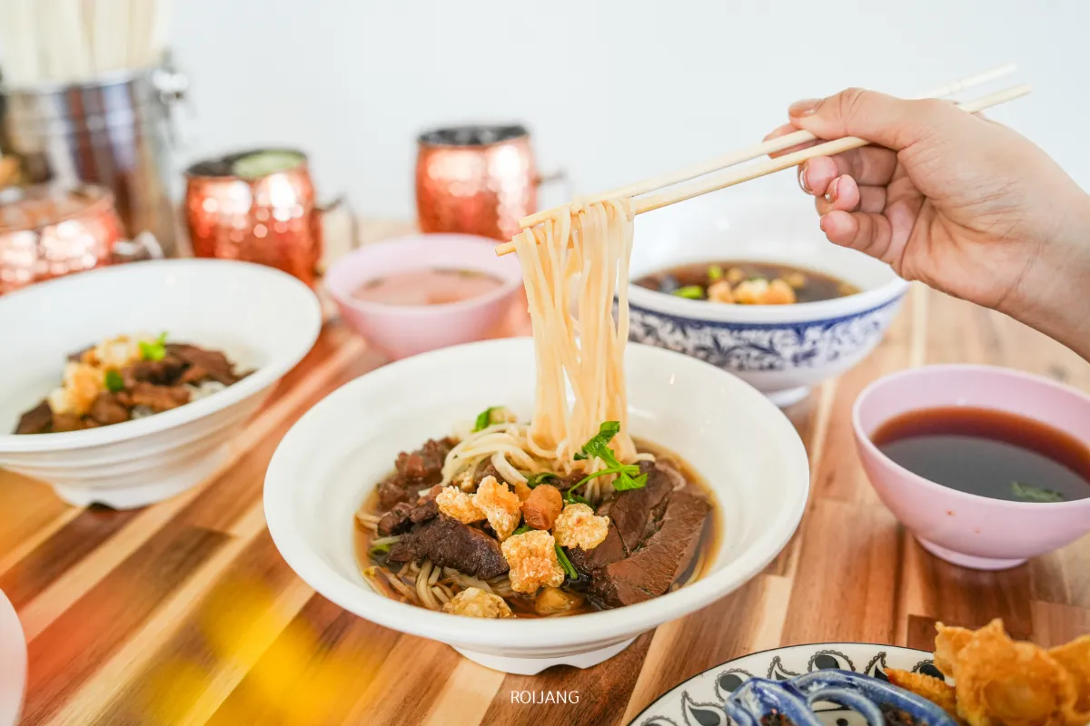 มือใช้ตะเกียบคีบเส้นก๋วยเตี๋ยวจากชามก๋วยเตี๋ยวเนื้อ ก๋งเส้นจันท์ โดยมีชามซุปและเครื่องเคียงอื่นๆ ล้อมรอบอยู่บนโต๊ะไม้.