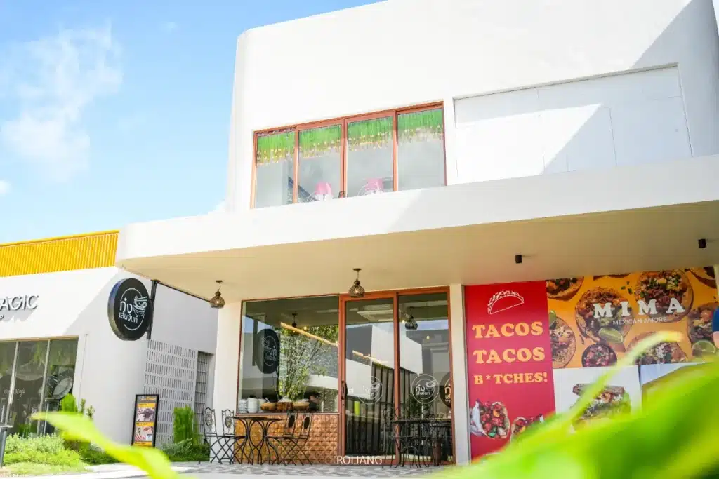 อาคารสีขาวสมัยใหม่ที่มีหน้าต่างบานใหญ่เป็นที่ตั้งของร้านอาหารชื่อ "มิมะ" ซึ่งมี ก๋วยเตี๋ยวเส้นจันท์, ที่นั่งกลางแจ้ง และป้ายโฆษณาที่สดใสสำหรับทาโก้.