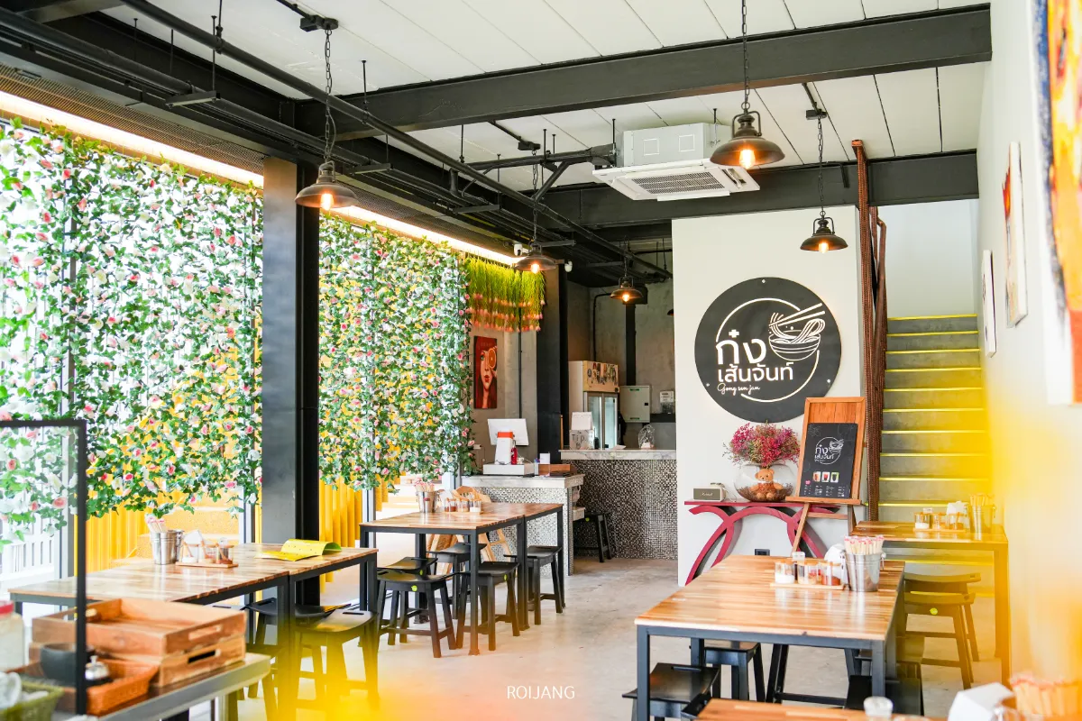 ภายในร้านอาหารที่สว่างและทันสมัย มีโต๊ะและเก้าอี้ไม้ ผ้าม่านลายดอกไม้ ศิลปะบนผนัง และเคาน์เตอร์ที่ด้านหลัง—เหมาะอย่างยิ่งสำหรับการเพลิดเพลินกับก๋วยเตี๋ยวเส้นจันท์.