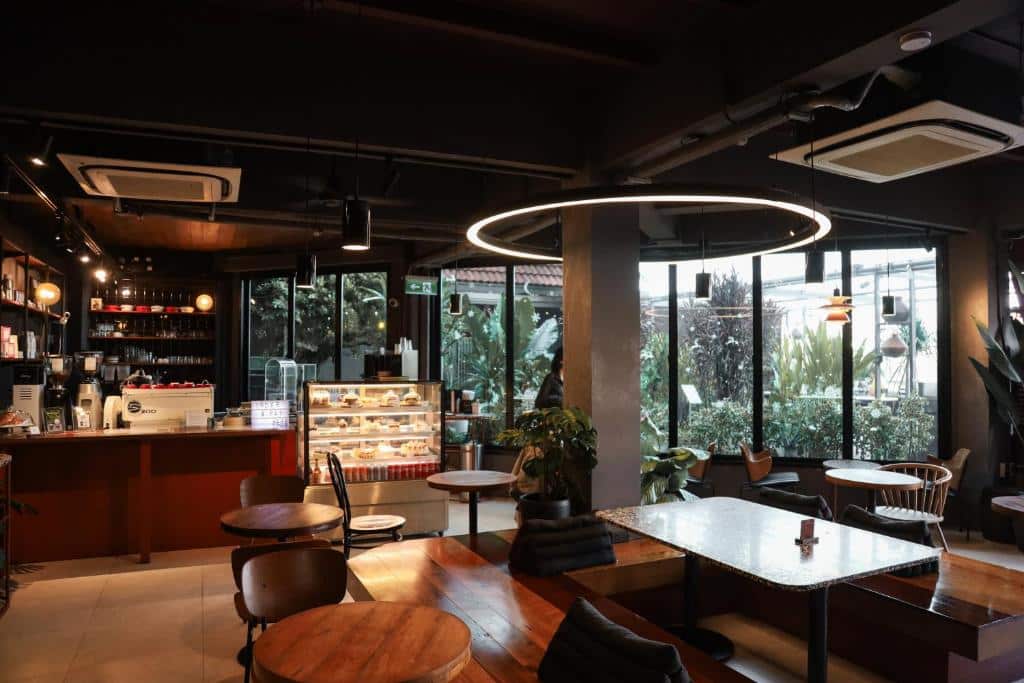 ภายในคาเฟ่สไตล์โมเดิร์นมีโต๊ะไม้ เก้าอี้ ตู้โชว์ขนมอบ และหน้าต่างบานใหญ่ที่ให้แสงธรรมชาติส่องเข้ามา โคมไฟทรงกลมติดเพดานมองเห็นได้เหนือพื้นที่นั่งเล่นตรงกลาง.