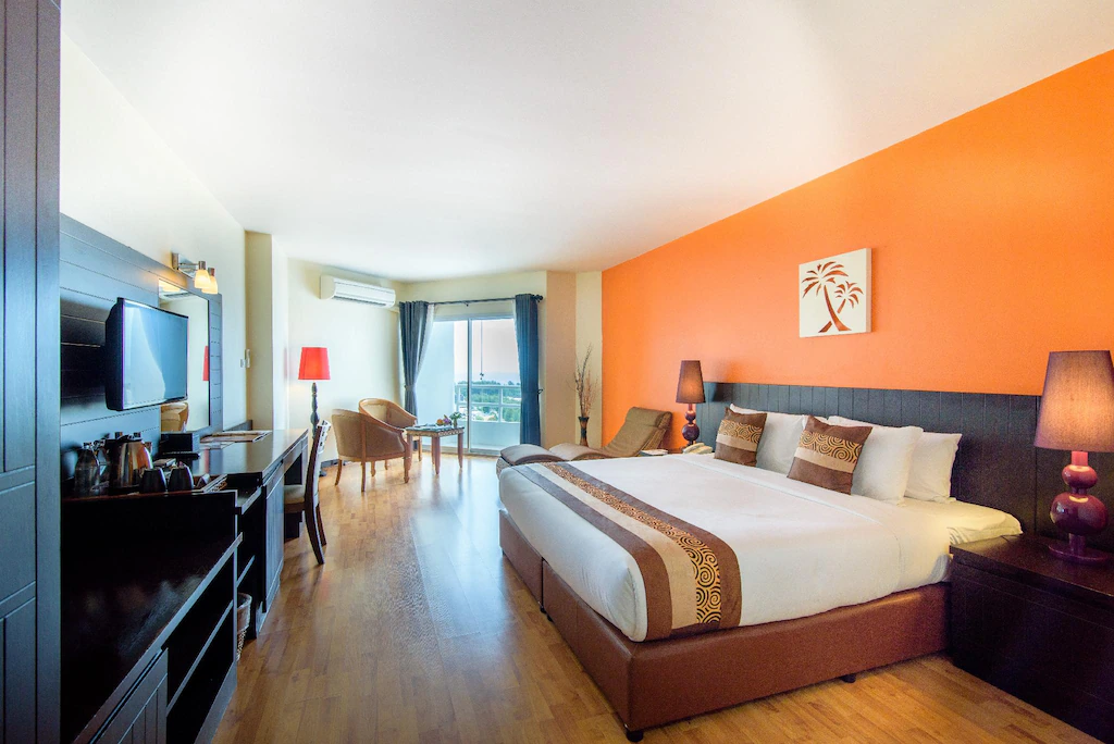 ห้องพักโรงแรมสมัยใหม่ที่ โรงแรมชะอำ พร้อมเตียงขนาดใหญ่ ผนังสีส้มเป็นจุดเด่น โคมไฟ โทรทัศน์ โต๊ะทำงาน และพื้นที่นั่งเล่นสบาย ๆ ริมหน้าต่างที่เต็มไปด้วยแสงธรรมชาติ.