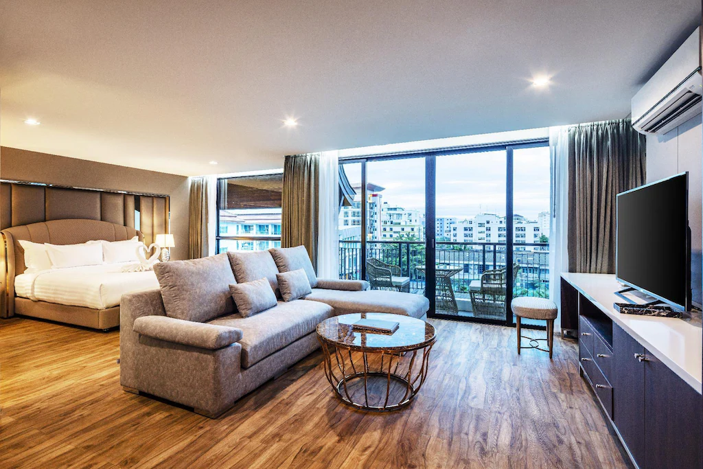 ห้องพักในโรงแรมทันสมัยพร้อมเตียงขนาดใหญ่ โซฟาสีเทา โต๊ะกาแฟทรงกลม ทีวีจอแบน และหน้าต่างบานใหญ่จากพื้นจรดเพดานที่เปิดสู่ระเบียงพร้อมทิวทัศน์เมือง.
