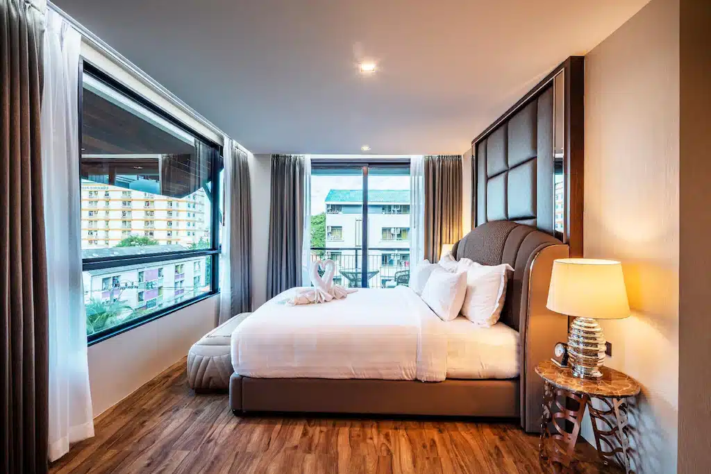 ห้องพักในโรงแรมที่ทันสมัยพร้อมเตียงขนาดใหญ่ ผ้าขนหนูพับลายหงส์ โคมไฟข้างเตียง พื้นไม้ และหน้าต่างบานใหญ่ที่มองเห็นอาคารภายนอก.
