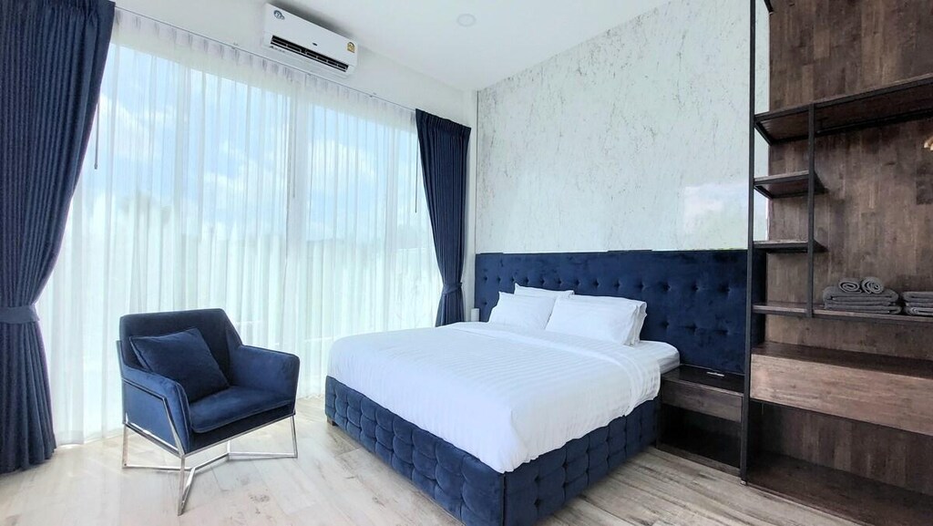ห้องนอนที่ทันสมัยพร้อมเตียงขนาดใหญ่ หัวเตียงบุด้วยเบาะสีน้ำเงิน เก้าอี้เท้าแขนสีน้ำเงิน ชั้นวางของแบบเปิดพร้อมผ้าขนหนู และหน้าต่างบานสูงจากพื้นจรดเพดานพร้อมผ้าม่านสีน้ำเงิน.