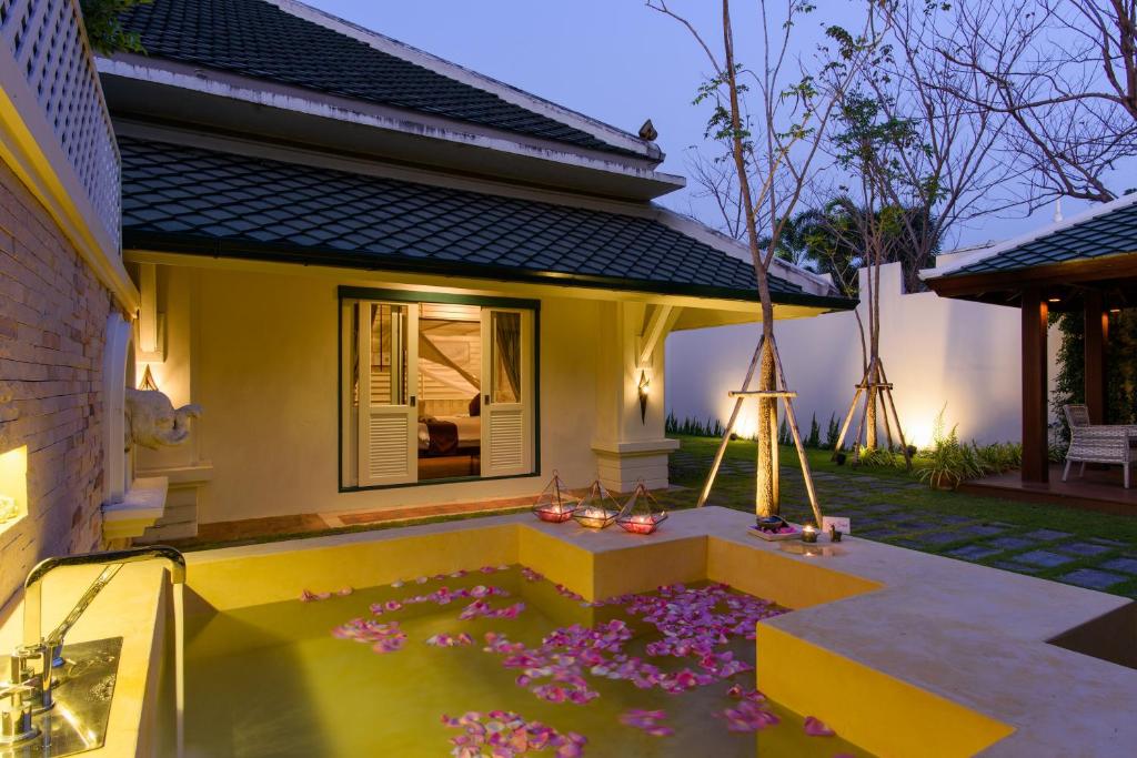 ห้องอาบน้ำกลางแจ้งที่เต็มไปด้วยน้ำและกลีบดอกไม้สีชมพูในลานบ้านที่มีบ้านหลังเล็ก หน้าต่างที่เปิดอยู่ และต้นไม้ในยามพลบค่ำ.