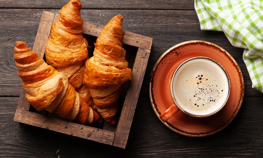ครัวซองต์สามชิ้นในกล่องไม้ข้างถ้วยกาแฟบนจานรองสีน้ำตาล พร้อมผ้าลายตารางสีเขียวและสีขาวในมุมห้อง เป็นภาพอาหารเช้าอันแสนอบอุ่นที่สมบูรณ์แบบก่อนที่คุณจะออกสำรวจ ที่เที่ยวสัตหีบ.