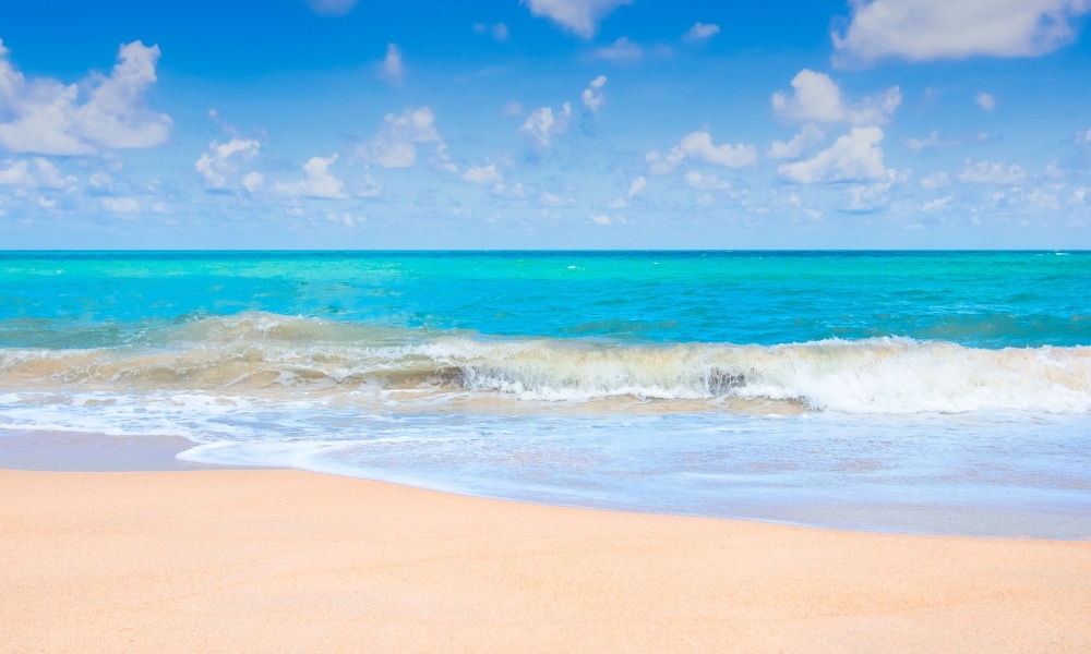 หาดทรายที่มีคลื่นอ่อนโยนและน้ำทะเลสีฟ้าครามภายใต้ท้องฟ้าสีครามและมีเมฆกระจาย เหมาะสำหรับผู้ที่มองหาที่เที่ยวสัตหีบหรือที่พักผ่อนที่พักผ่อนสัตหีบ.