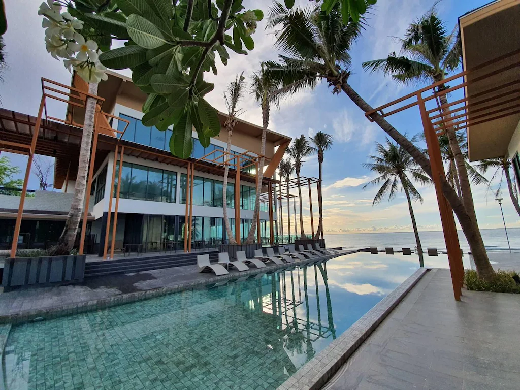 อาคารริมชายหาดสมัยใหม่พร้อมหน้าต่างกระจกขนาดใหญ่ ต้นปาล์ม และสระว่ายน้ำกลางแจ้งพร้อมเก้าอี้อาบแดด เหมาะสำหรับการพักผ่อนที่ที่พักชะอำ ชมวิวท้องฟ้าและเมฆ.