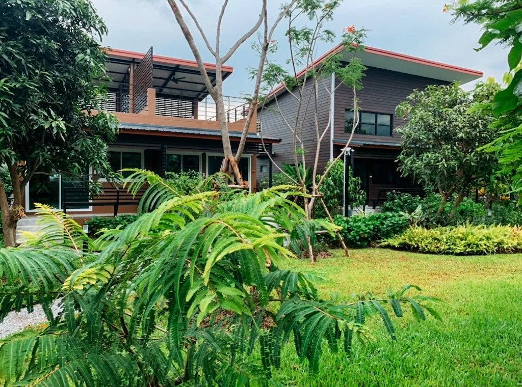 บ้านสองชั้นสไตล์โมเดิร์นพร้อมหน้าต่างบานใหญ่และสวนที่ประดับด้วยต้นไม้สีเขียว ต้นไม้ และสนามหญ้าที่ได้รับการดูแลอย่างดีภายใต้ท้องฟ้าที่มีเมฆมาก.