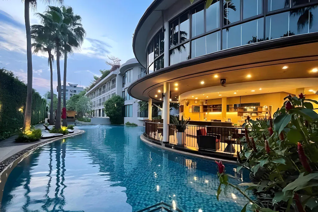 โรงแรมทันสมัยพร้อมด้วยผนังโค้งและหน้าต่างบานใหญ่ที่มองเห็นสระว่ายน้ำกลางแจ้ง ล้อมรอบด้วยต้นปาล์มและพืชพรรณเขียวขจีในยามพลบค่ำ—เหมาะสำหรับผู้ที่มองหาที่พักชะอำในบรรยากาศเงียบสงบ.