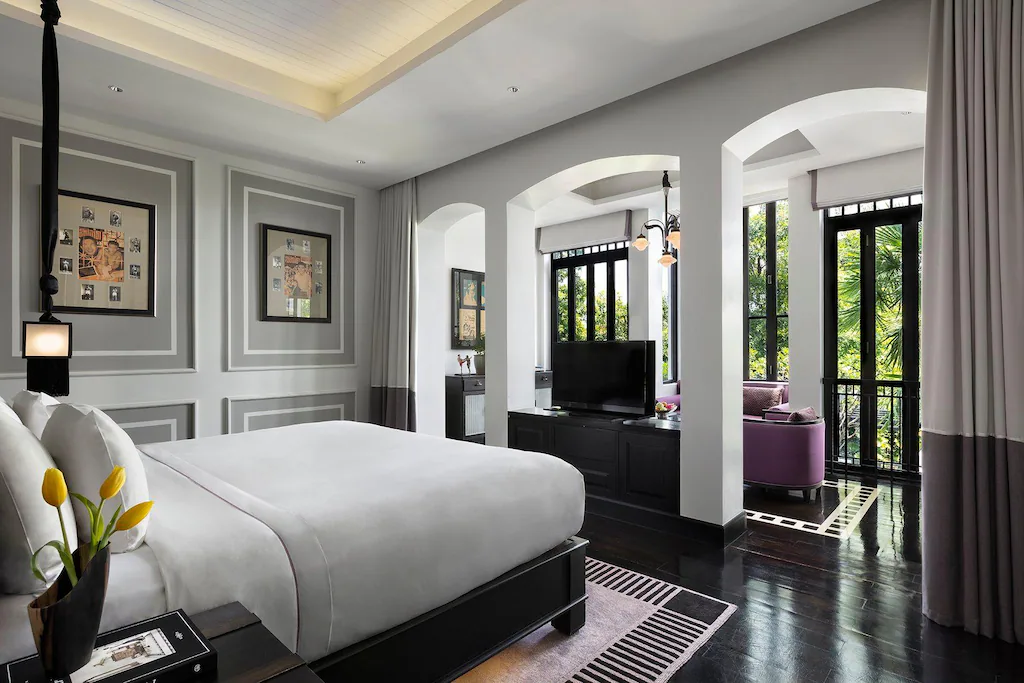 ห้องพักในโรงแรมที่ทันสมัยพร้อมเตียงนอนที่จัดอย่างประณีต พื้นสีดำ ภาพศิลปะแบบใส่กรอบบนผนังสีเทา ทีวีบนตู้ และหน้าต่างบานใหญ่ที่มองเห็นทิวทัศน์ของต้นไม้สีเขียว.