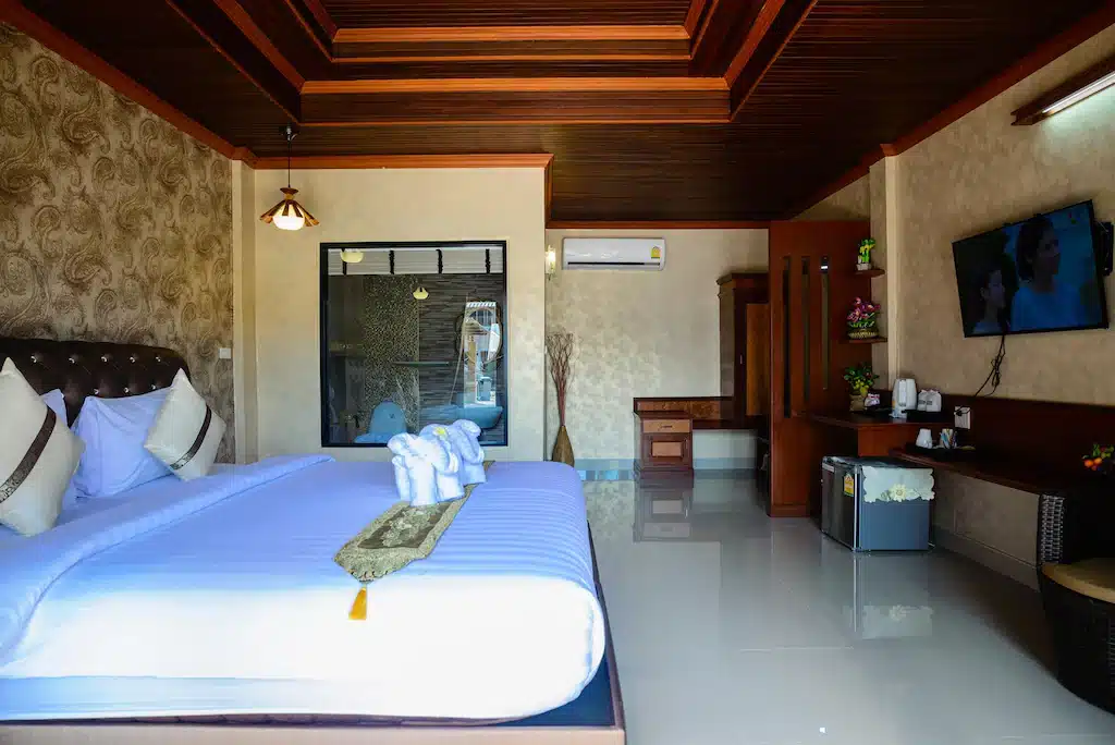 ห้องพักในโรงแรมที่มีเตียงขนาดใหญ่ งานศิลปะเกี่ยวกับผ้าเช็ดตัวแบบพับ  ที่พักสตูลแนวธรรมชาติ ทีวีจอแบน โต๊ะทำงาน ตู้เย็นขนาดเล็ก และห้องน้ำผนังกระจกเป็นพื้นหลัง เหมาะสำหรับผู้ที่กำลังมองหาสตูลแนวธรรมชาติพร้อมความสะดวกสบายที่ทันสมัย.