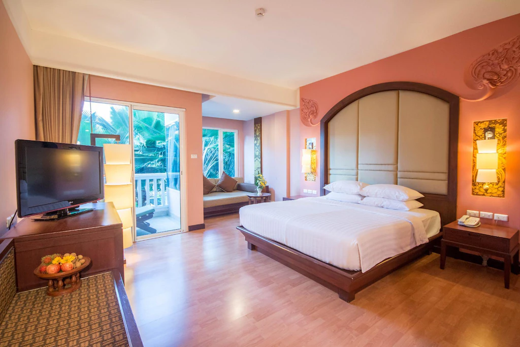 ห้องพักโรงแรมที่ โรงแรมชะอำ พร้อมเตียงขนาดใหญ่ โคมไฟติดผนัง ทีวี โซฟาข้างหน้าต่าง โต๊ะเล็กพร้อมผลไม้ และระเบียงที่มองเห็นวิวต้นไม้เขียวขจี—เหมาะสำหรับการพักผ่อนของคุณที่ชะอำ.