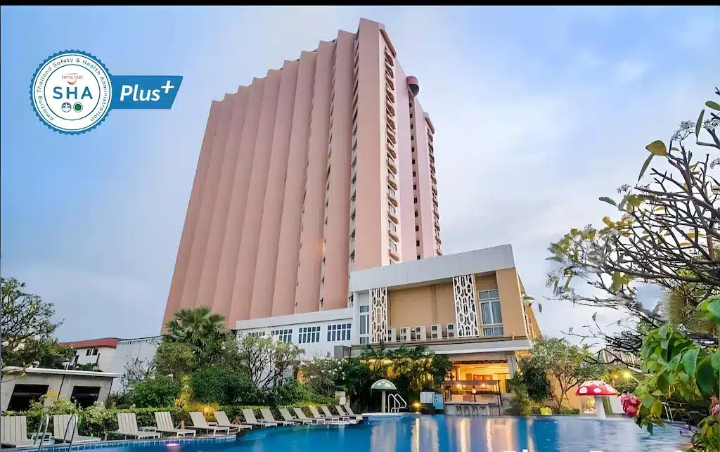 โรงแรมสีชมพูสูงหลายชั้น พร้อมเก้าอี้ริมสระว่ายน้ำ และต้นไม้ในเบื้องหน้า เหมาะสำหรับผู้ที่ต้องการที่พักชะอำราคาถูก โลโก้การรับรอง SHA Plus+ อยู่ที่มุมบนซ้าย.