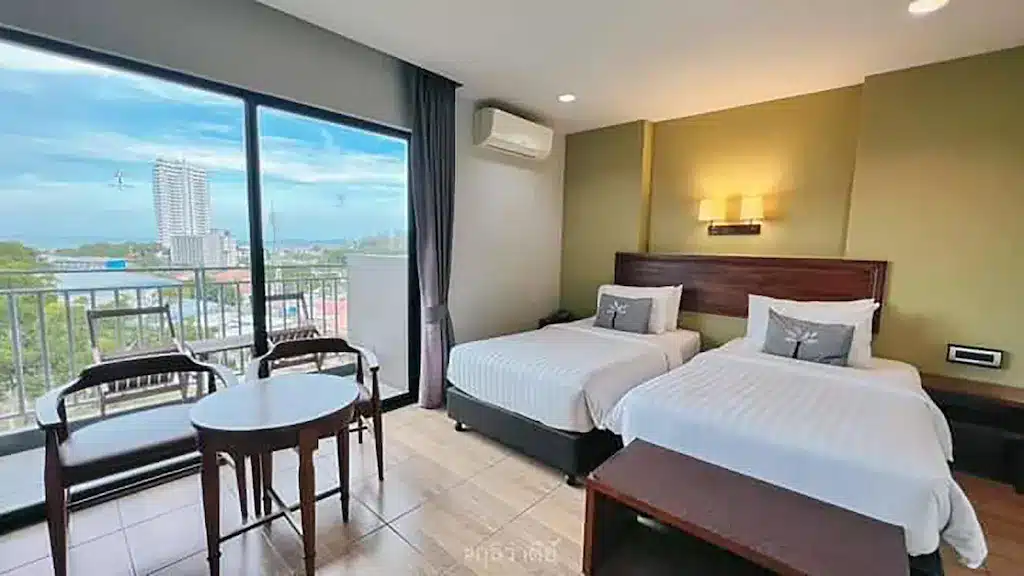 ห้องพักในโรงแรมพร้อมเตียงเดี่ยว 2 เตียง โต๊ะและเก้าอี้ขนาดเล็กริมหน้าต่างบานใหญ่ที่มองเห็นอาคารและต้นไม้ เพลิดเพลินกับการตกแต่งที่เรียบง่ายและทันสมัย เหมาะสำหรับผู้ที่กำลังมองหาร้านอาหารสัตหีบหลังจากเที่ยวชมที่เที่ยวสัตหีบในบริเวณใกล้เคียง.