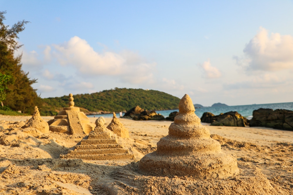 ประติมากรรมทรายในรูปร่างของเจดีย์เรียงรายอยู่บนชายหาดทรายที่ ที่เที่ยวสัตหีบ ตั้งอยู่ริมทะเล โขดหิน และเนินเขาที่ปกคลุมไปด้วยต้นไม้ใต้ท้องฟ้าที่มีเมฆบางส่วน เป็นฉากที่เชื้อเชิญให้มาผ่อนคลายและสำรวจ.