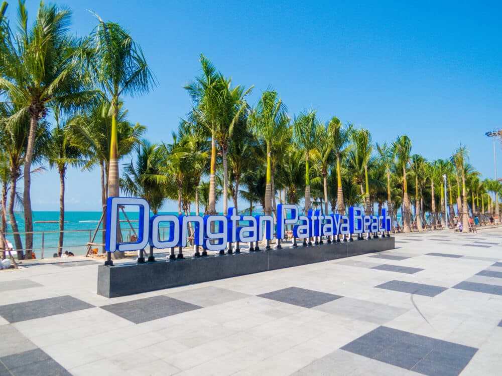 ทางเดินที่เรียงรายไปด้วยต้นปาล์มริมทะเลมีป้ายขนาดใหญ่เขียนว่า "หาดดงตาลพัทยา" ใต้ท้องฟ้าสีฟ้าใส ทำให้ที่นี่เป็นสถานที่ยอดนิยมสำหรับผู้ที่ต้องการไปเที่ยวสัตหีบหรือหาที่พักผ่อนสัตหีบ.