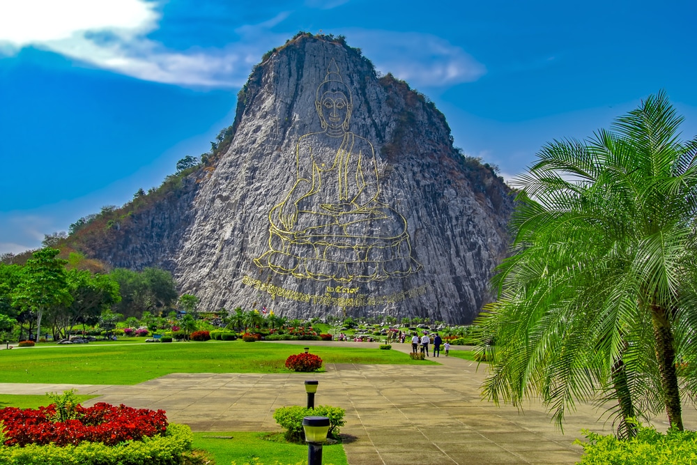 ที่เที่ยวสัตหีบ พระพุทธรูปทองคำขนาดใหญ่แกะสลักไว้บนหน้าเนินหิน ล้อมรอบด้วยสนามหญ้าสีเขียว ต้นปาล์ม ดอกไม้ และท้องฟ้าสีครามสดใส.
