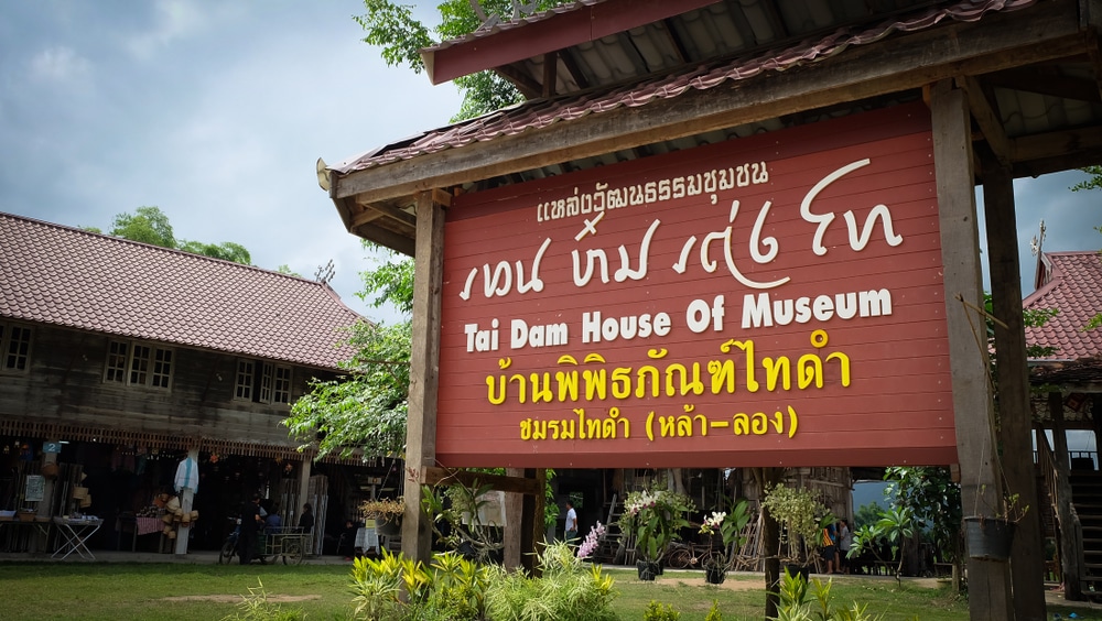 ป้ายทางเข้าบ้านไทดำของพิพิธภัณฑ์ ซึ่งเป็นสถานที่ท่องเที่ยวอันดับต้นๆ เที่ยวเลย  ของเลย พร้อมอาคารไม้แบบดั้งเดิมและนักท่องเที่ยวในพื้นหลังภายใต้ท้องฟ้าที่มีเมฆครึ้ม.