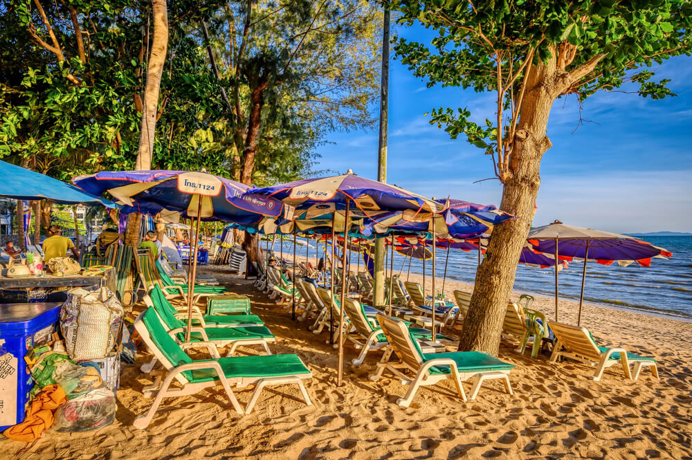 เก้าอี้เลานจ์สีเขียวเรียงรายพร้อมร่มสีน้ำเงินและสีม่วงตั้งอยู่บนชายหาดทรายที่สถานที่ท่องเที่ยวสัตหีบยอดนิยม โดยมีต้นไม้ให้ร่มเงาและมองเห็นทะเลเป็นฉากหลัง.