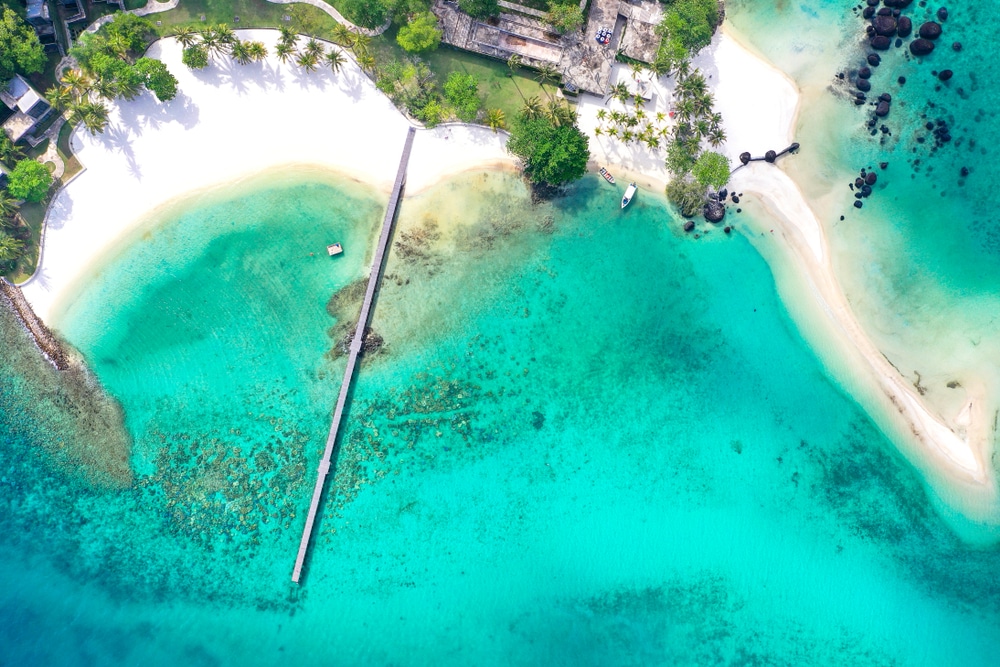 มุมมองทางอากาศของชายหาดเขตร้อนแห่งหนึ่งในเที่ยวสัตหีบที่มีทรายขาว น้ำทะเลสีฟ้าใส ท่าเรือยาว ต้นปาล์ม และอาคารหลายหลังใกล้ชายฝั่ง เหมาะสำหรับการพักผ่อนอย่างผ่อนคลาย.