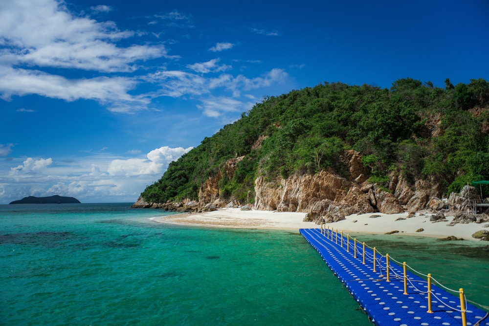 ท่าเรือลอยน้ำสีฟ้าทอดยาวไปสู่หาดทรายที่มีน้ำทะเลสีฟ้าครามใส และไหล่เขาหินเขียวขจีใต้ท้องฟ้าสีครามสดใส เหมาะสำหรับผู้ที่มองหาที่พักผ่อนสัตหีบหรือสำรวจที่เที่ยวสัตหีบ.