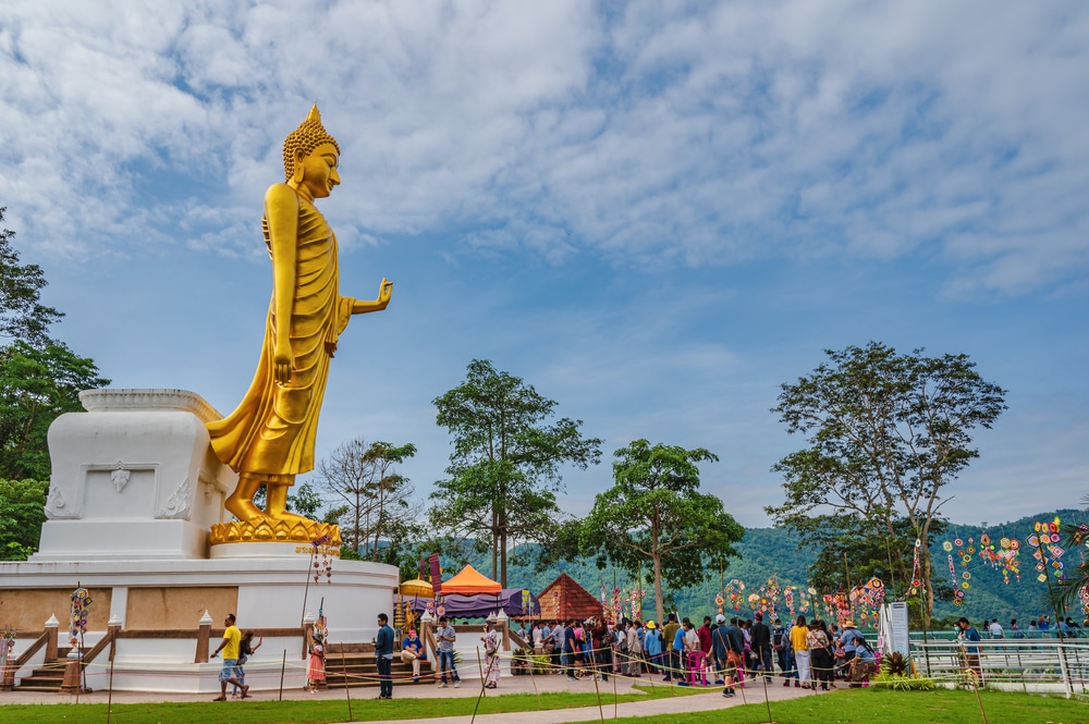 พระพุทธรูปทองคำขนาดใหญ่ตั้งอยู่ในวัดกลางแจ้ง ที่ผู้คนมารวมตัวกันอยู่รอบต้นไม้และของตกแต่ง ท่ามกลางท้องฟ้า ที่เที่ยวเลย ที่มีเมฆบางส่วนปกคลุม — สถานที่ท่องเที่ยวที่ต้องไปเยือนในจังหวัดเลยสำหรับผู้ที่ต้องการสำรวจบริเวณนี้.