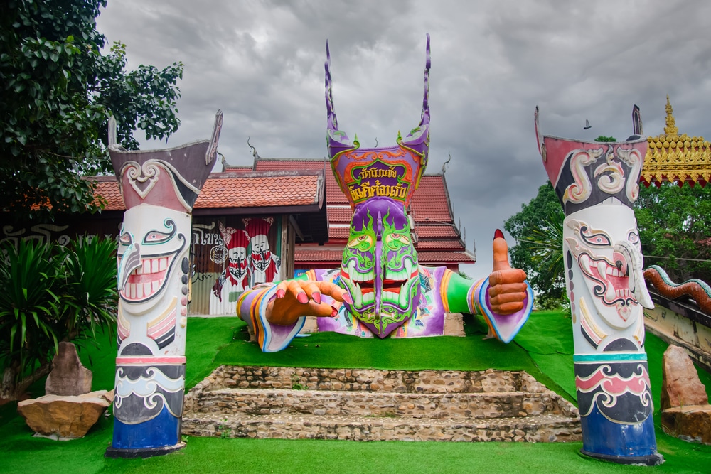 รูปปั้นสีสันสดใสที่มีหน้ากากขนาด ที่เที่ยวเลย ใหญ่และแขนที่เหยียดออกกำลังยกนิ้วโป้งให้ โดยมีเสาที่ทาสีสองต้นขนาบข้าง อยู่ด้านหน้าของอาคารไทยแบบดั้งเดิม—เหมาะสำหรับผู้ที่ต้องการสำรวจสถานที่ท่องเที่ยวในเลย.