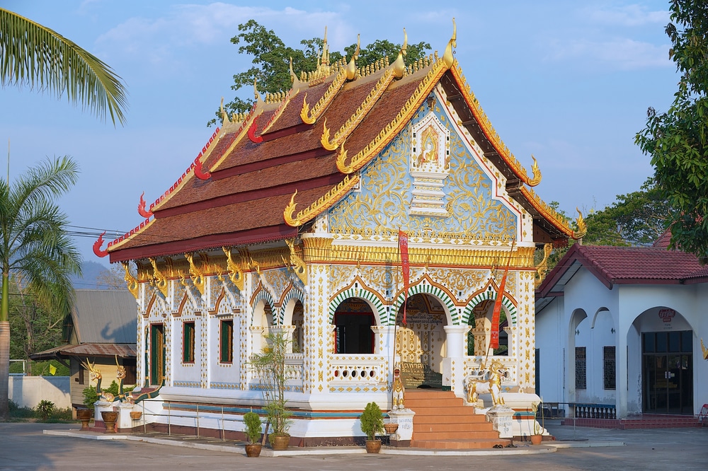วัดพุทธไทยที่ประดับประดาด้วยทองคำอย่างวิจิตร มีหลังคาสีแดง  ที่เที่ยวเลย และลวดลายที่ซับซ้อนบนผนังด้านหน้า ตั้งอยู่ท่ามกลางต้นไม้และอาคารใกล้เคียงในวันที่ท้องฟ้าแจ่มใส—เหมาะสำหรับผู้ที่ต้องการสถานที่ท่องเที่ยวเพื่อสำรวจวัฒนธรรมท้องถิ่น.