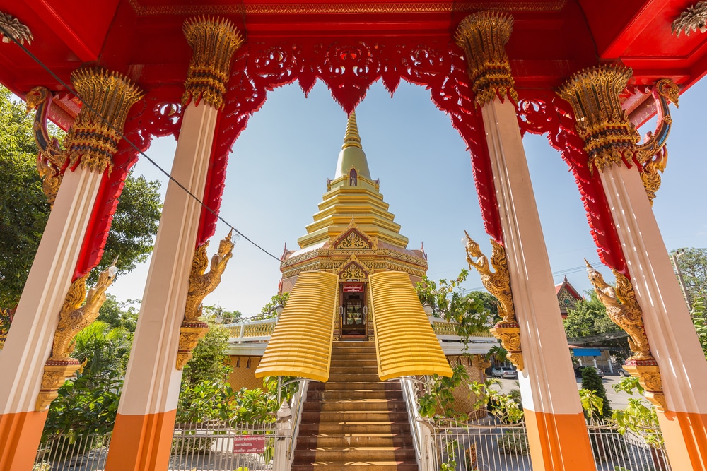 เจดีย์สีทองที่ประดับด้วยรายละเอียดสีแดงและสีทองอันวิจิตร มองผ่านซุ้มประตูที่ตกแต่งอย่างวิจิตรบรรจง โดยมีขั้นบันไดทอดยาวไปจนถึงทางเข้า สถานที่อันเงียบสงบที่เหมาะสำหรับการพักผ่อนในสัตหีบหรือค้นพบสถานที่ใหม่ๆ ที่เที่ยวสัตหีบ.
