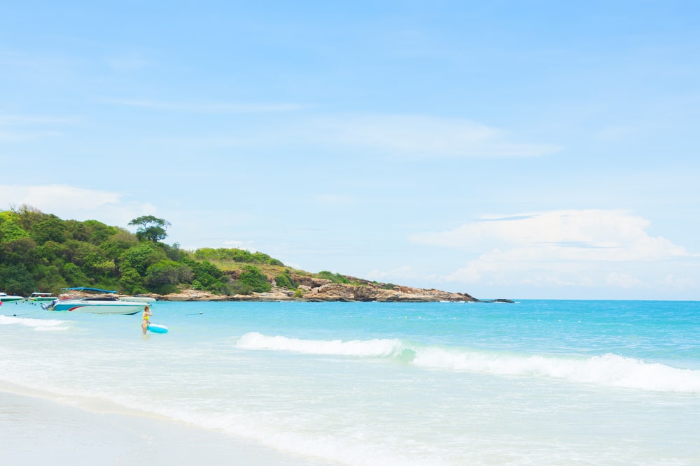 หาดทรายที่มีคลื่นอ่อนโยน น้ำทะเลสีฟ้าคราม คนเดินถือกระดานโต้คลื่น และเนินเขาเขียวขจีใต้ท้องฟ้าสีฟ้าใส เหมาะสำหรับเที่ยวสัตหีบและการพักผ่อนสัตหีบยอดนิยมสำหรับรักทะเล.