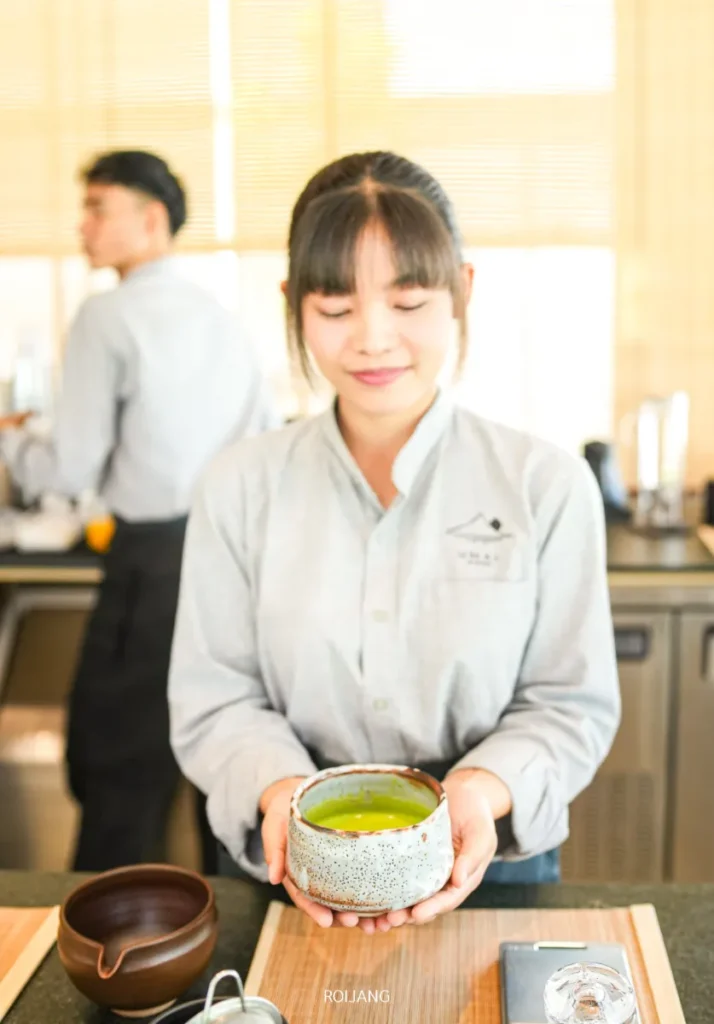 ผู้หญิงในชุดเครื่องแบบสีเทาอ่อนถือชามชาเขียวที่ร้านชา UMAI Teahouse ยืนอยู่หลังเคาน์เตอร์ที่มีอุปกรณ์ชงชา; มีผู้ชายคนหนึ่งทำงานอยู่เบื้องหลัง.