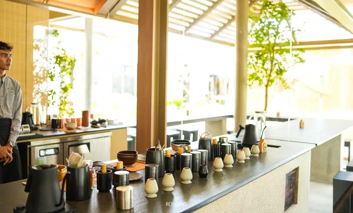 บาร์กาแฟสมัยใหม่แบบเปิดโล่ง มีถ้วย แก้ว และอุปกรณ์ชงกาแฟหลากหลายชนิดจัดวางอย่างเป็นระเบียบบนเคาน์เตอร์ พนักงานคนหนึ่งยืนอยู่ทางซ้าย สร้างบรรยากาศที่ชวนให้นึกถึง UMAI Teahouse.