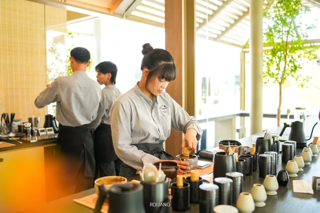 บาริสต้าในเสื้อสีเทาเตรียมกาแฟที่เคาน์เตอร์ของ UMAI Teahouse ในขณะที่พนักงานอีกสองคนพูดคุยกันอยู่เบื้องหลังของคาเฟ่ที่สว่างไสว.