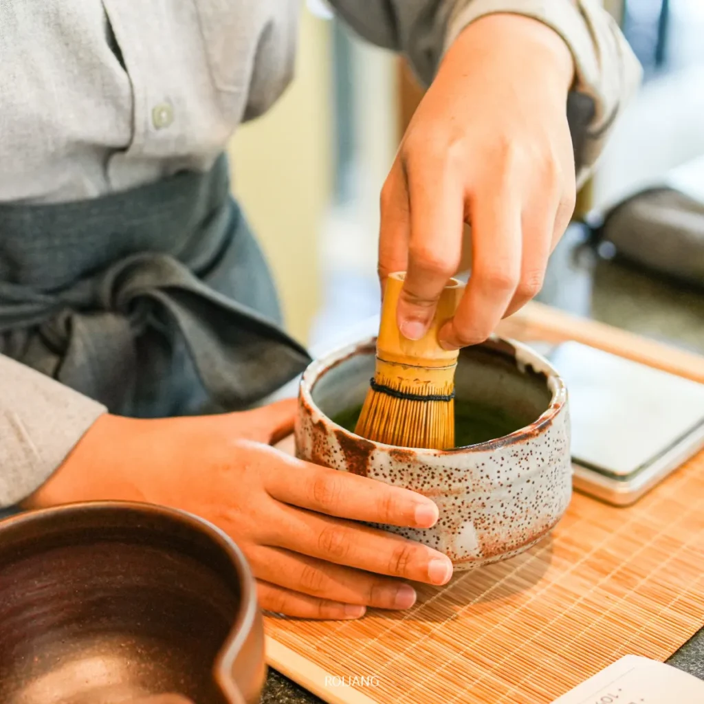 บุคคลหนึ่งใช้ตะกร้อไม้ไผ่ตีชามัทฉะในถ้วยเซรามิกบนเสื่อไม้ไผ่ที่ร้านชา UMAI Teahouse.
