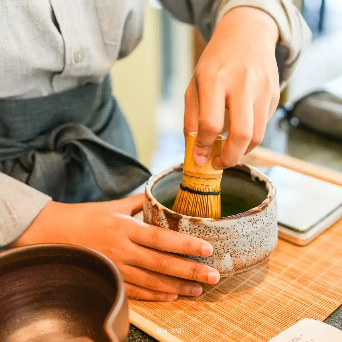 บุคคลหนึ่งใช้ตะกร้อไม้ไผ่ตีชามัทฉะในถ้วยเซรามิกบนเสื่อไม้ไผ่ที่ร้านชา UMAI Teahouse.
