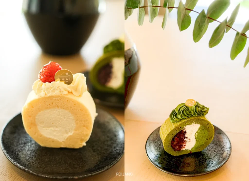 เค้กโรลสองชิ้นจาก UMAI Teahouse บนจานสีดำ: ชิ้นหนึ่งเป็นวานิลลาพร้อมครีมและสตรอเบอร์รี่ อีกชิ้นเป็นมัทฉะพร้อมครีมและไส้ถั่วแดง วางบนโต๊ะสีอ่อนที่มีต้นไม้อยู่ด้านบน.