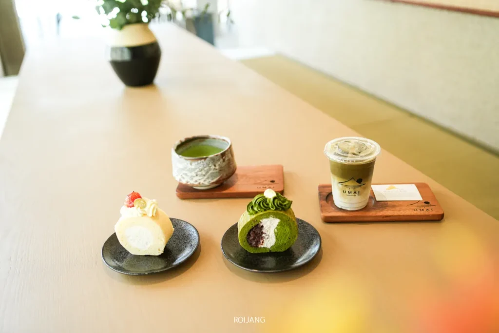 สองจานของขนมหวาน, ถ้วยชาเขียวร้อน, และเครื่องดื่มเย็นถูกจัดวางบนโต๊ะไม้ในบรรยากาศสมัยใหม่ของ UMAI Teahouse.