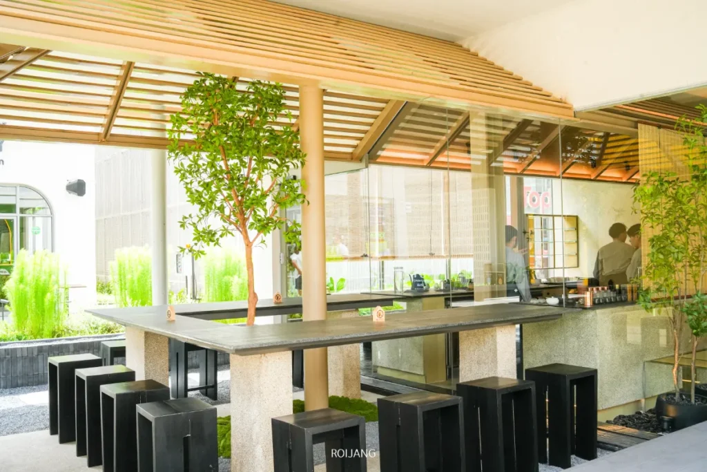 พื้นที่นั่งเล่นกลางแจ้งสไตล์โมเดิร์นที่ UMAI Teahouse ประกอบด้วยโต๊ะยาว เก้าอี้สตูลสีดำ ต้นไม้ในกระถาง และหลังคาบางส่วน โดยมีผู้คนกำลังทำงานอยู่หลังเคาน์เตอร์ในฉากหลัง.