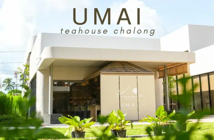 อาคารภายนอกสมัยใหม่พร้อมหน้าต่างกระจกบานใหญ่และป้ายที่เขียนว่า "UMAI Teahouse Chalong" ล้อมรอบด้วยพืชพรรณเขียวขจีภายใต้ท้องฟ้าแจ่มใส สัมผัสกับบรรยากาศที่เชิญชวนของ UMAI Teahouse ในสถานที่ที่เงียบสงบนี้.