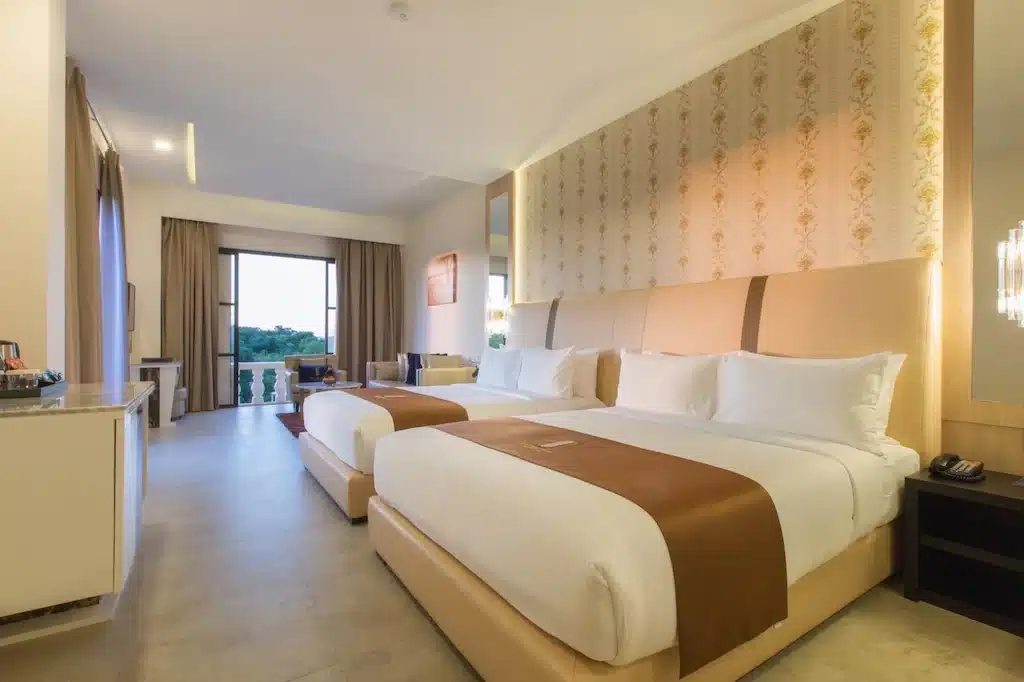 ห้องพักโรงแรมที่ โรงแรมเขาใหญ่ พร้อมเตียงใหญ่สองเตียง ผ้าปูเตียงสีน้ำตาล ผนังตกแต่งลวดลาย โต๊ะทำงาน มุมนั่งเล่น และระเบียงที่มองเห็นวิวทิวทัศน์สี ที่พักเขาใหญ่ เขียวขจี.