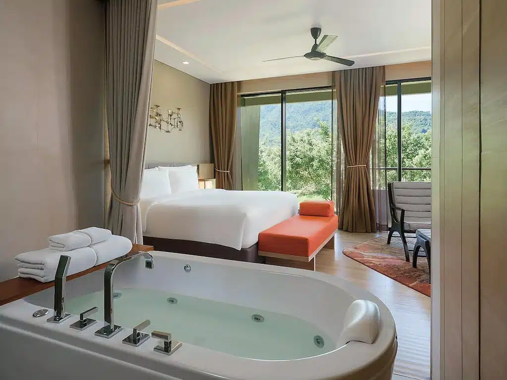 ห้องพักโรงแรมสมัยใหม่พร้อมเตียงขนาดใหญ่ ม้านั่งสีส้ม พื้นที่นั่งเล่น และอ่างอาบน้ำแบบผ่อนคลาย; ประตูบานกระจกเปิดออกสู่ระเบียงที่มองเห็นต้นไม้และภูเขา—เหมาะสำหรับผู้ที่มองหา ที่พักเขาใหญ่ หรือที่พักเขาใหญ่ราคาประหยัด.