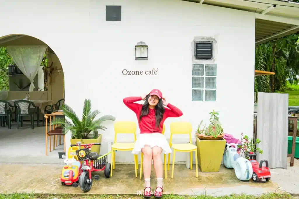 ผู้หญิงคนหนึ่งสวมเสื้อสีแดงและหมวก นั่งอยู่บนเก้าอี้สีเหลือง หน้าร้านโอโซนคาเฟ่ ซึ่งเป็นอาคารสีขาว ล้อมรอบด้วยของเล่นรถยนต์และต้นไม้.