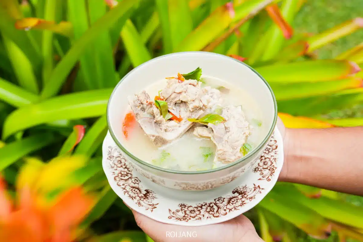 บุคคลหนึ่งถือชามซุปใสที่มีซี่โครงหมู สมุนไพร และผัก ตั้งอยู่ที่ร้านโอโซนคาเฟ่ โดยมีฉากหลังเป็นใบไม้สีเขียวและส้ม.