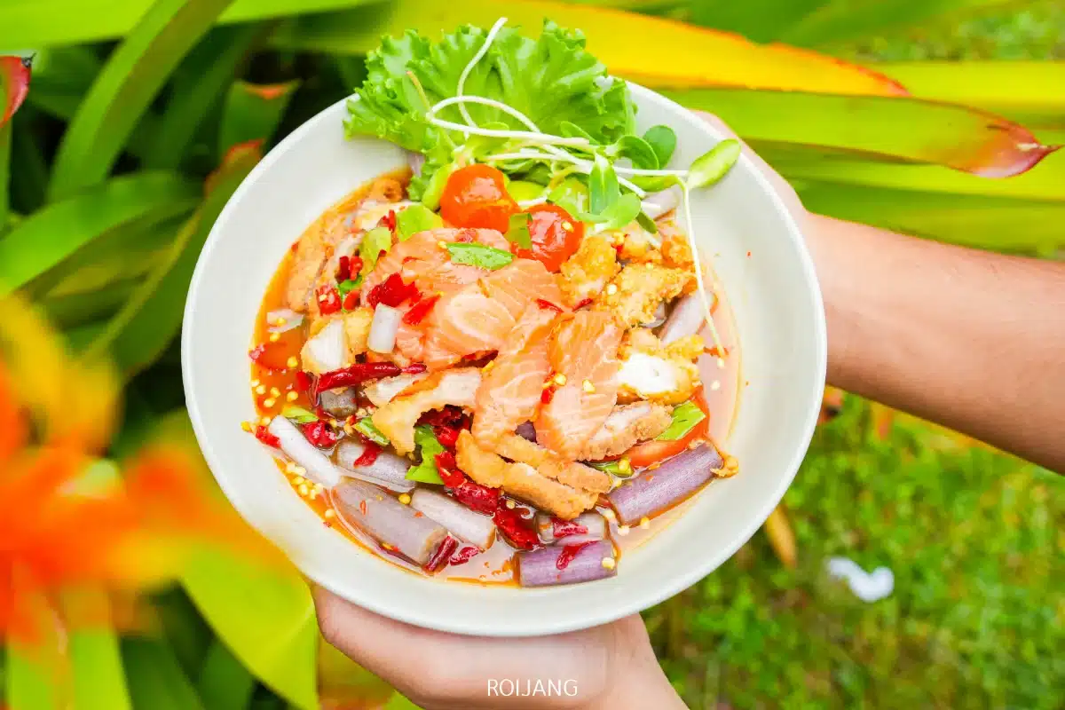 ชามสีขาวของสลัดไทยรสเผ็ดจาก Ozone Cafe พร้อมด้วยแซลมอนหั่นบาง หัวหอม ผักกาดหอม มะเขือเทศเชอร์รี่ พริกแห้ง และสมุนไพรต่างๆ วางอยู่กลางแจ้งท่ามกลางใบไม้สีเขียว.